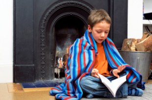 Boy in blanket on floor reading book by fireplace